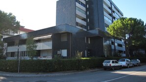 A modern multi-story building with a flat roof and large windows. There are several parked cars in front of the building. - Modern Flat at Grado Pineta With Terrace (Grado)