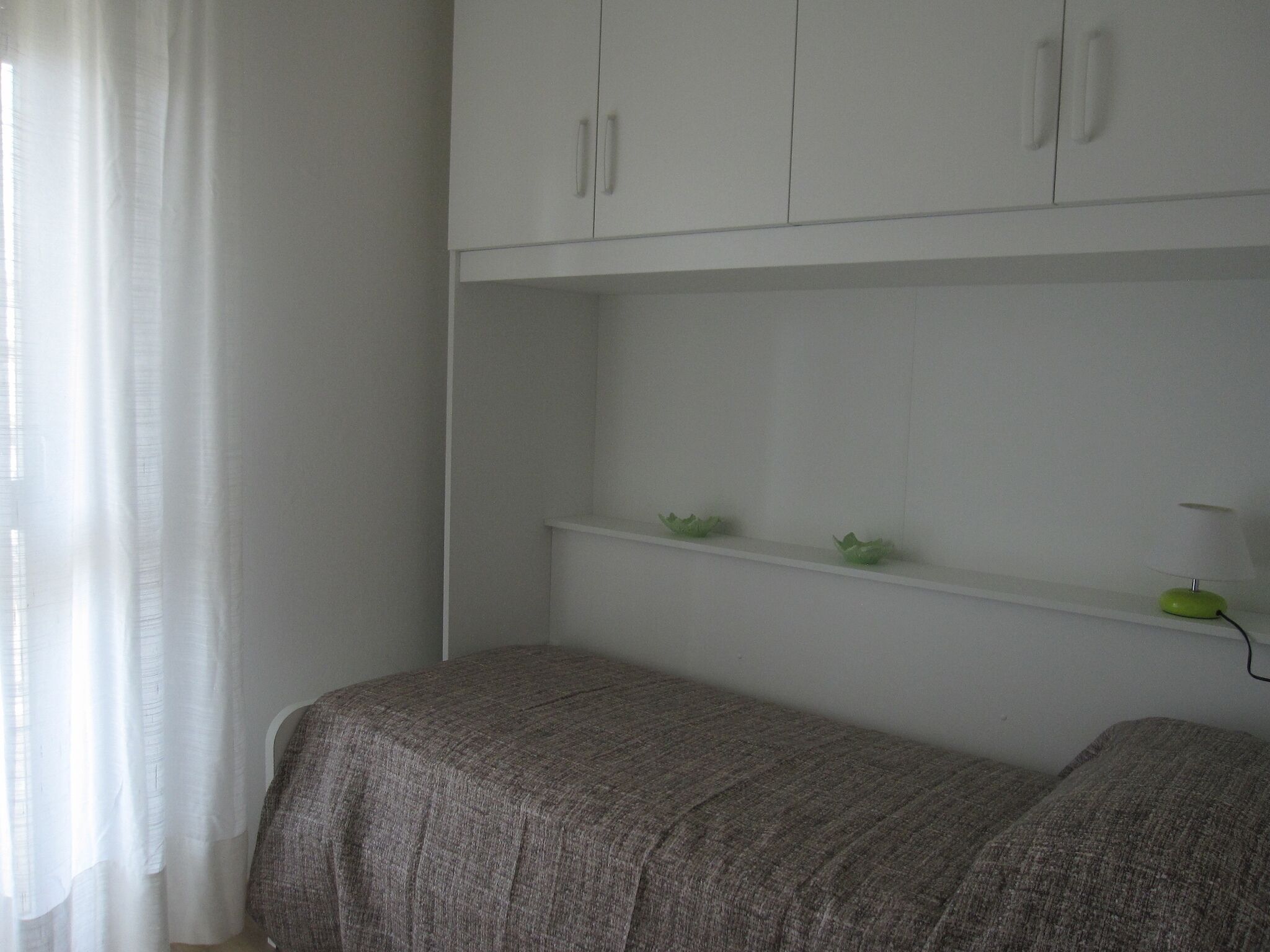 A small, neatly organized room with a bed, a white wardrobe, and a shelf with decorative items.