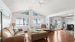 Ein geräumiges Wohnzimmer mit einer braunen Ledersofa, einem gestreiften Ottomanen und einem Essbereich mit Tisch und Stühlen. Große Fenster sorgen für viel natürliches Licht und Ausblick ins Freie.
