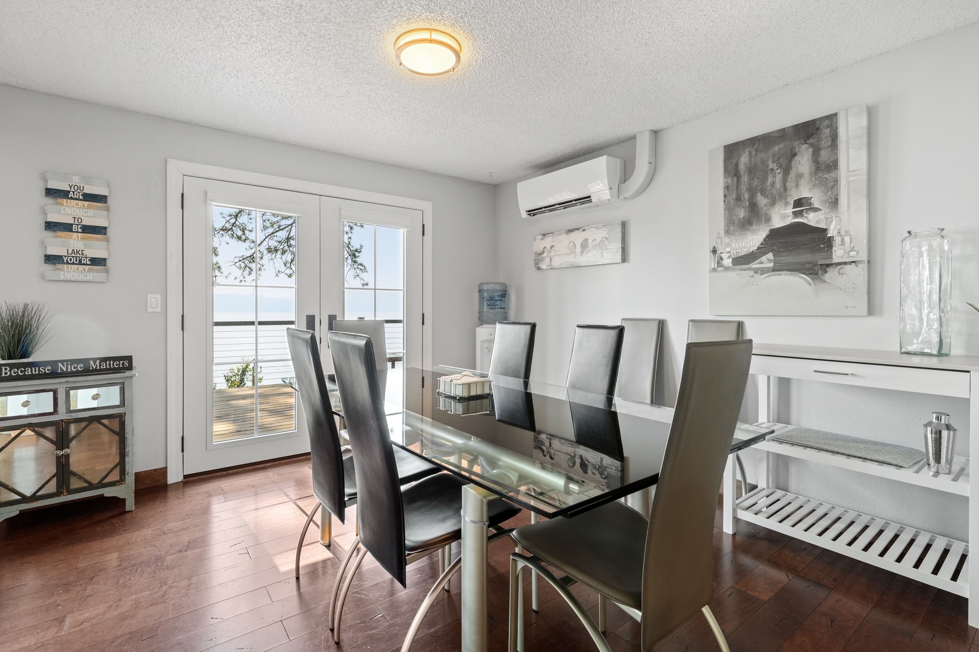 Un espace repas moderne avec une table en verre et des chaises noires, une console blanche agrémentée d’objets décoratifs, et une vue sur l’extérieur à travers une porte vitrée.