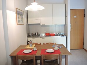A dining area with a wooden table set for two, a kitchen in the background, and a door to the right. - Flat in Residence With Swimming Pool and Balcony (San Michele al Tagliamento)
