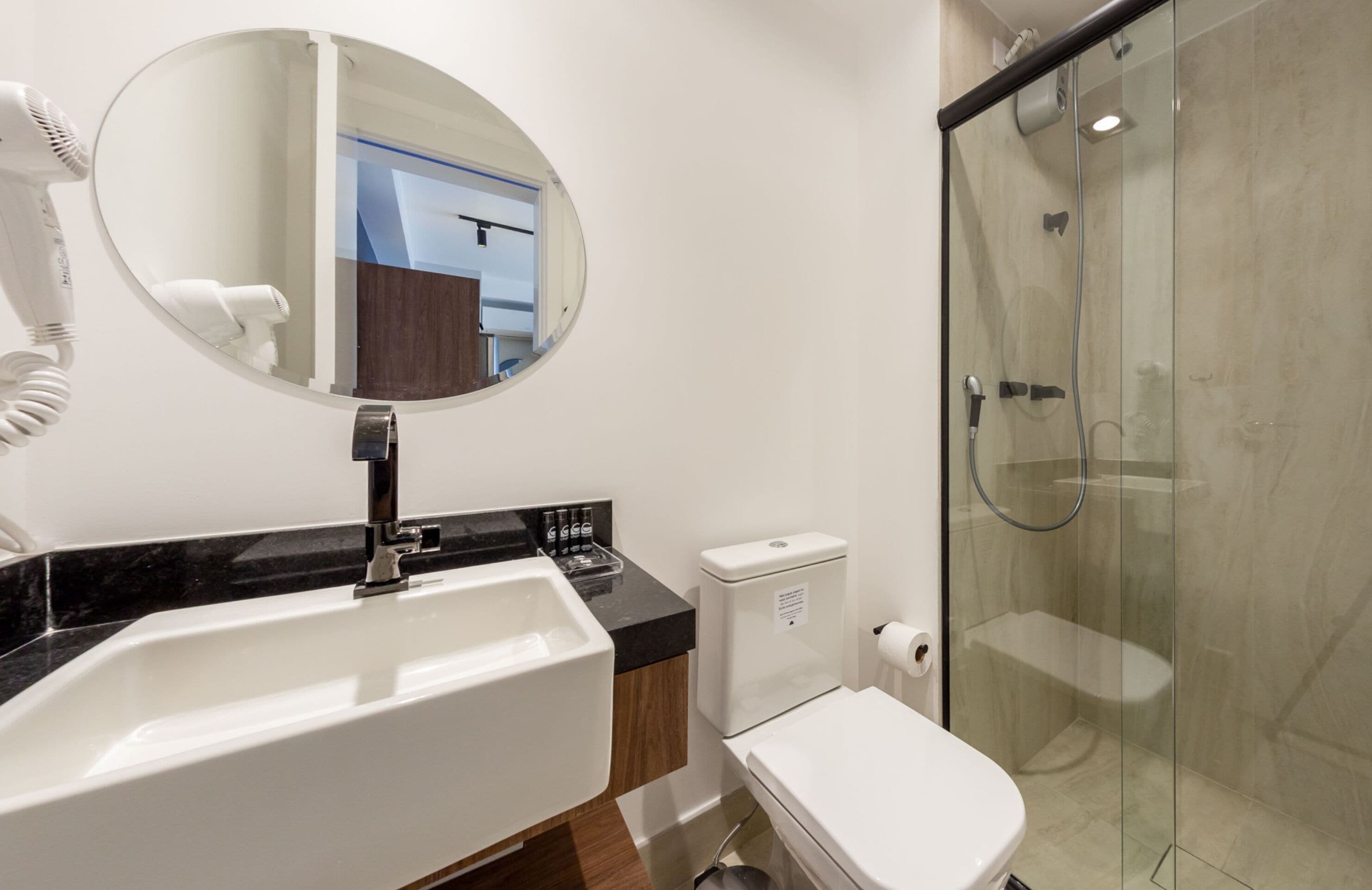 A modern bathroom with a white sink, a round mirror, a hairdryer, a toilet, and a glass-enclosed shower.