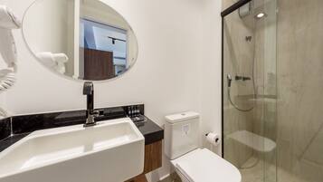 A modern bathroom with a white sink, a round mirror, a hairdryer, a toilet, and a glass-enclosed shower.