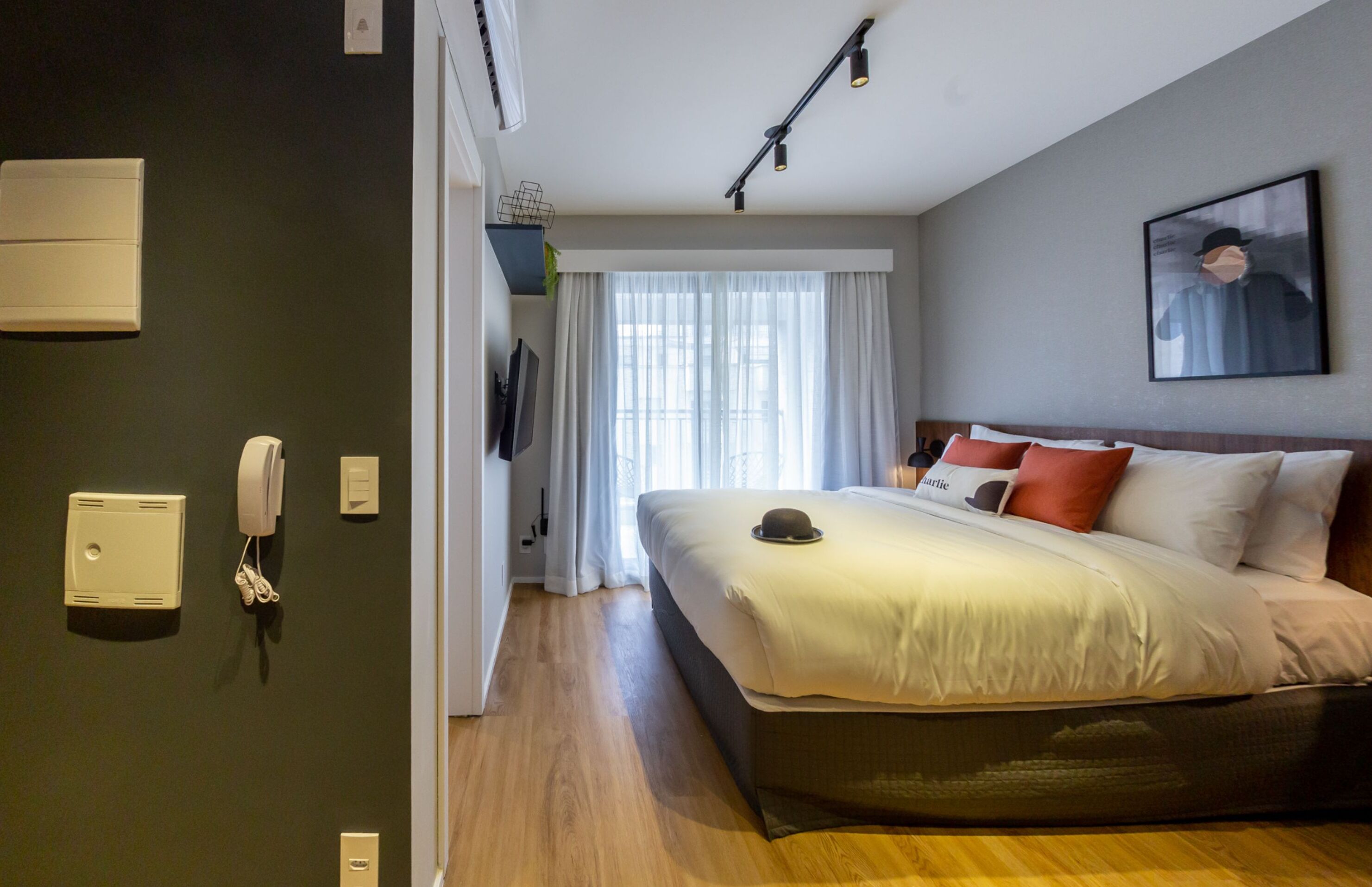 A modern hotel room with a large bed, a flat-screen TV mounted on the wall, a telephone, and a window with sheer curtains.