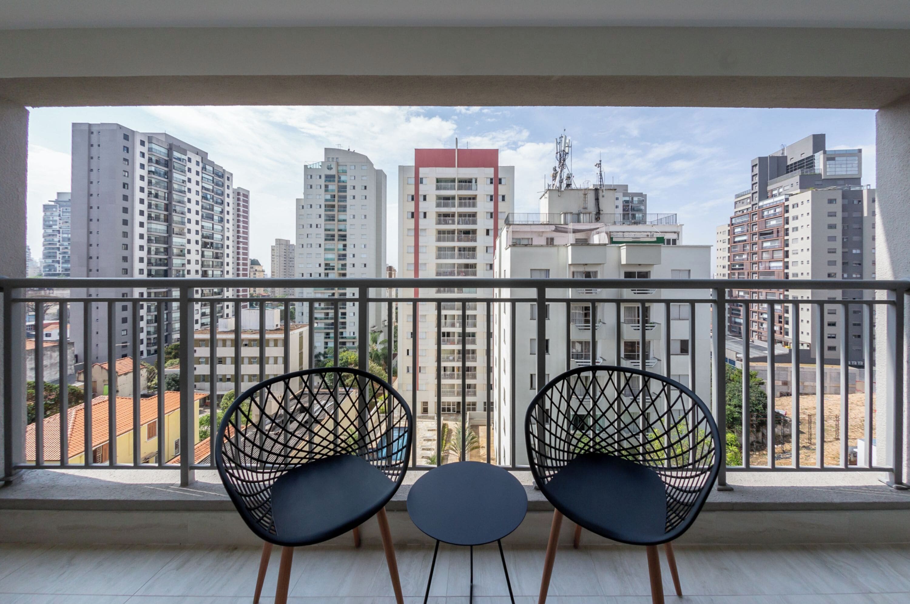 Ein Balkon mit zwei schwarzen Stühlen und einem kleinen Tisch, mit Blick auf eine Stadtlandschaft mit modernen Gebäuden.