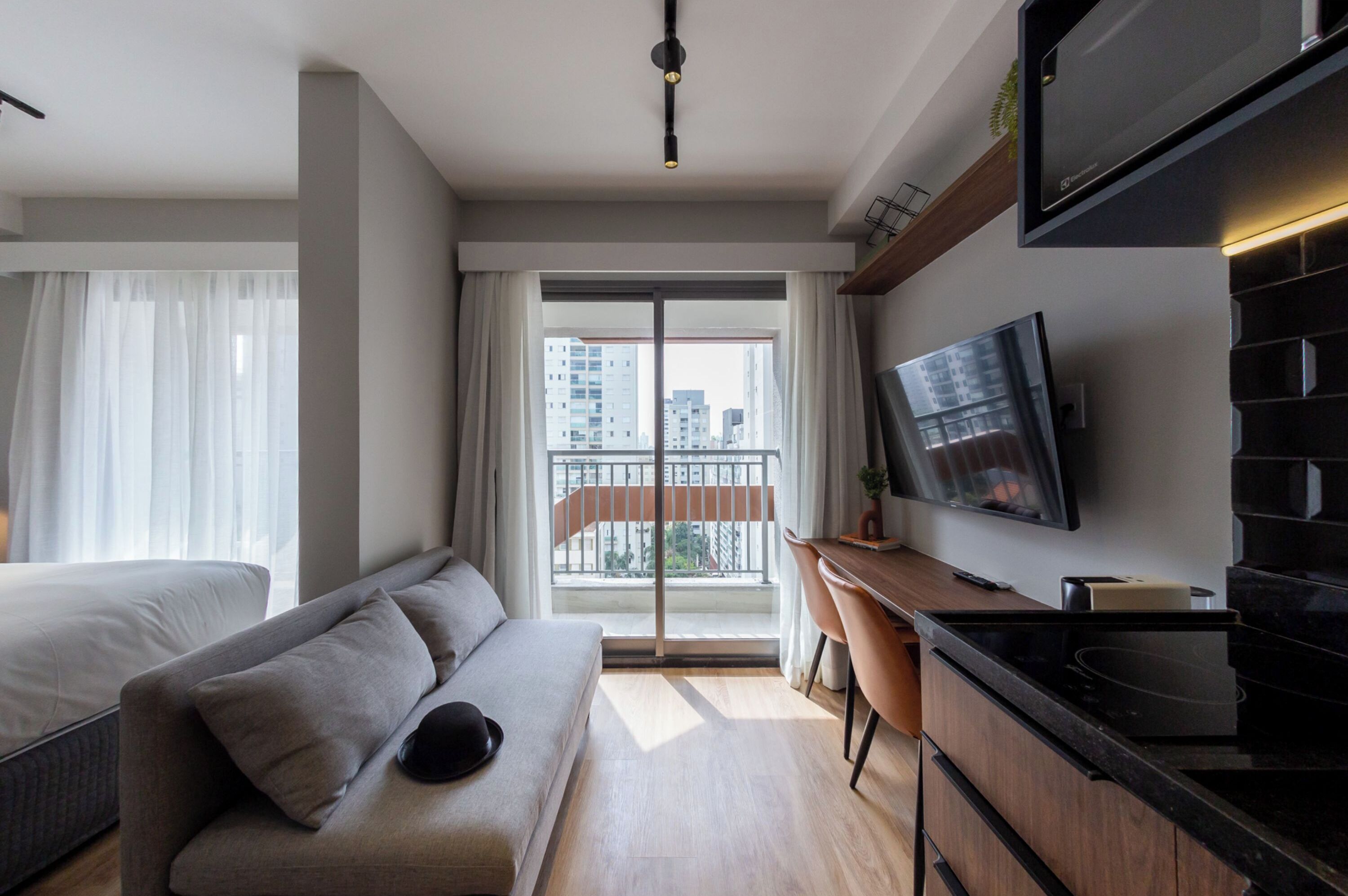 A modern hotel room with a sofa bed, a desk, a flat-screen TV, and a view of the city through large windows.