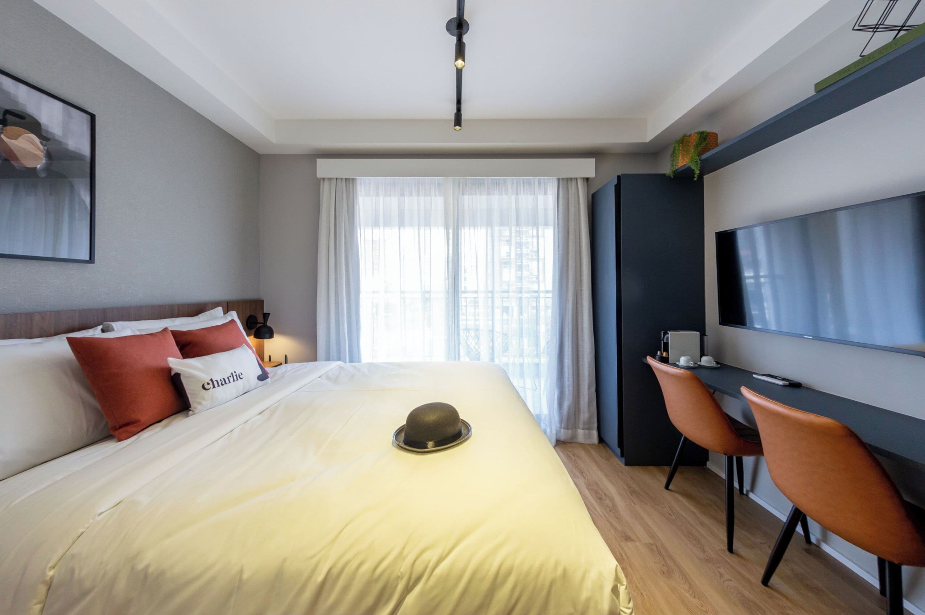 A modern hotel room with a large bed, a desk with a chair, a flat-screen TV, and a large window with sheer curtains.