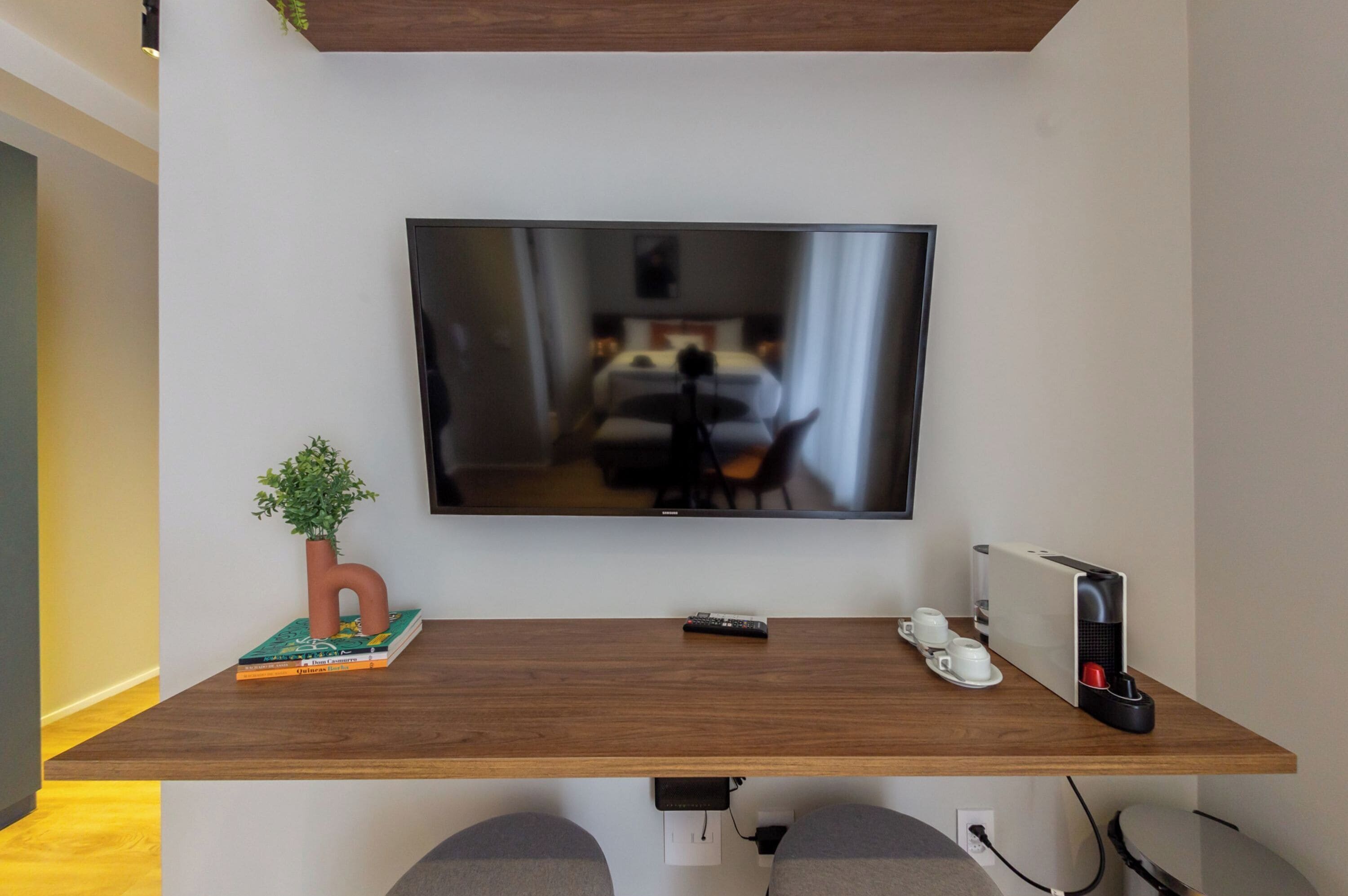Une chambre d’hôtel équipée d’un téléviseur à écran plat fixé au mur, d’un bureau en bois agrémenté d’une plante, d’une télécommande, d’une machine à café et d’un ordinateur portable.