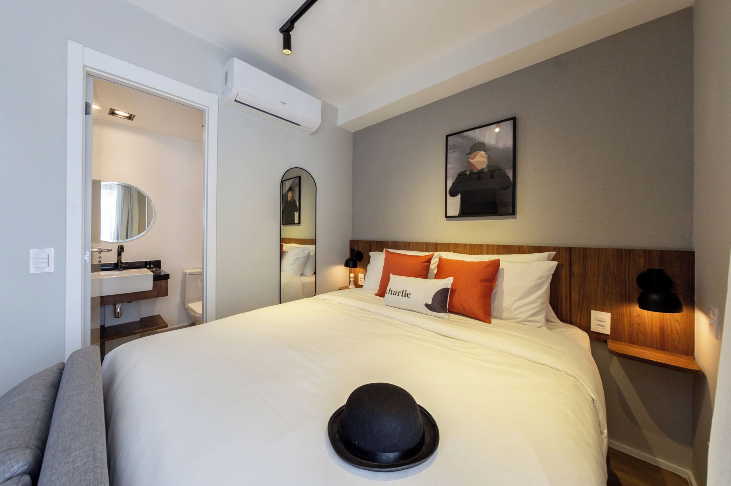 A hotel room with a bed, a bedside table, a mirror, a wall-mounted picture, and a small shelf with a lamp.