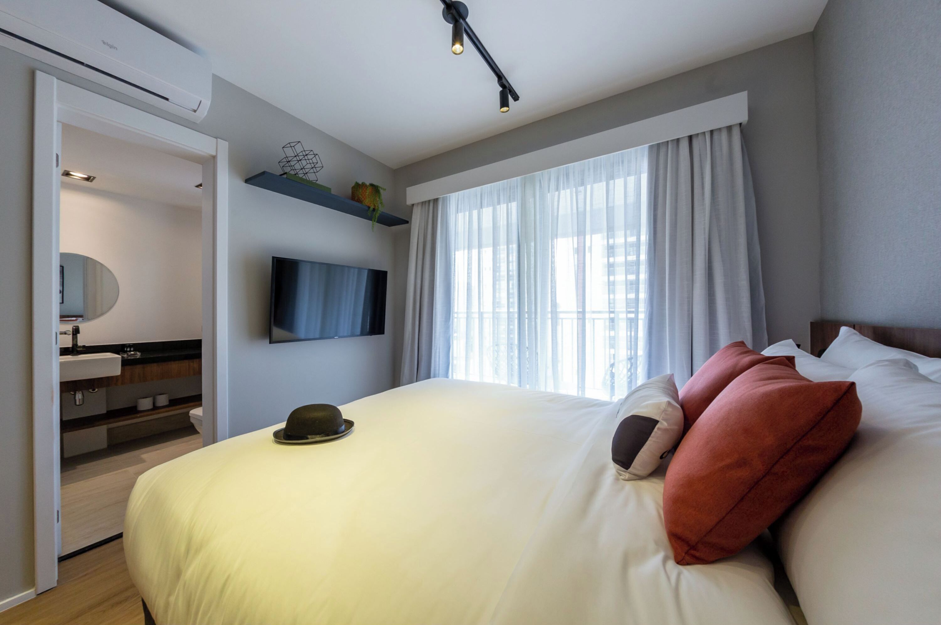 Et moderne hotelværelse med en stor seng, et fladskærms-tv på væggen, en indbygget toiletbord med et spejl og et vindue med lette gardiner.
