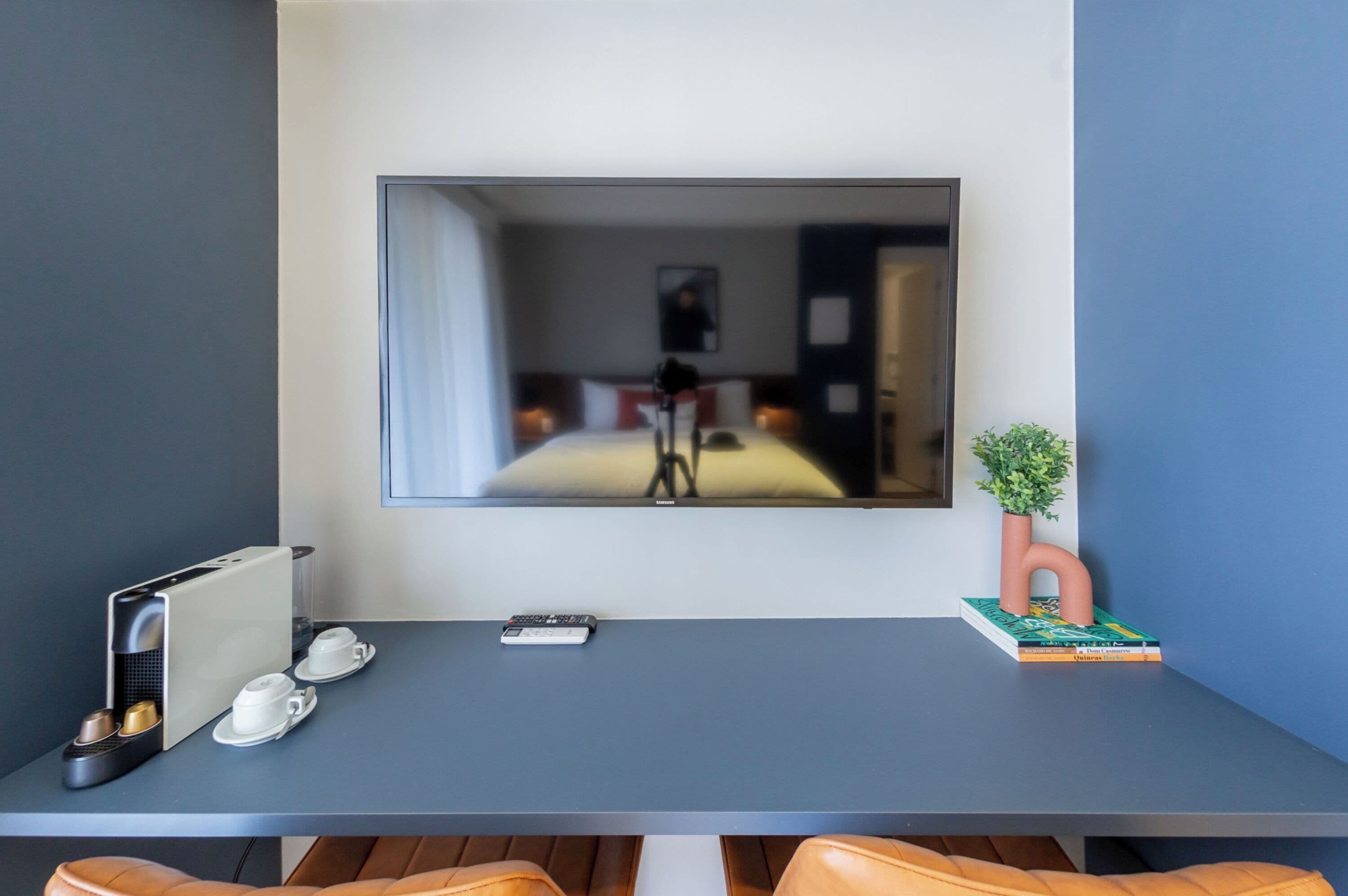 Ein modernes Hotelzimmer mit großem Flachbildfernseher an der Wand, einem kleinen Tisch mit Fernbedienung, einer Kaffeemaschine und einem Blumentopf.