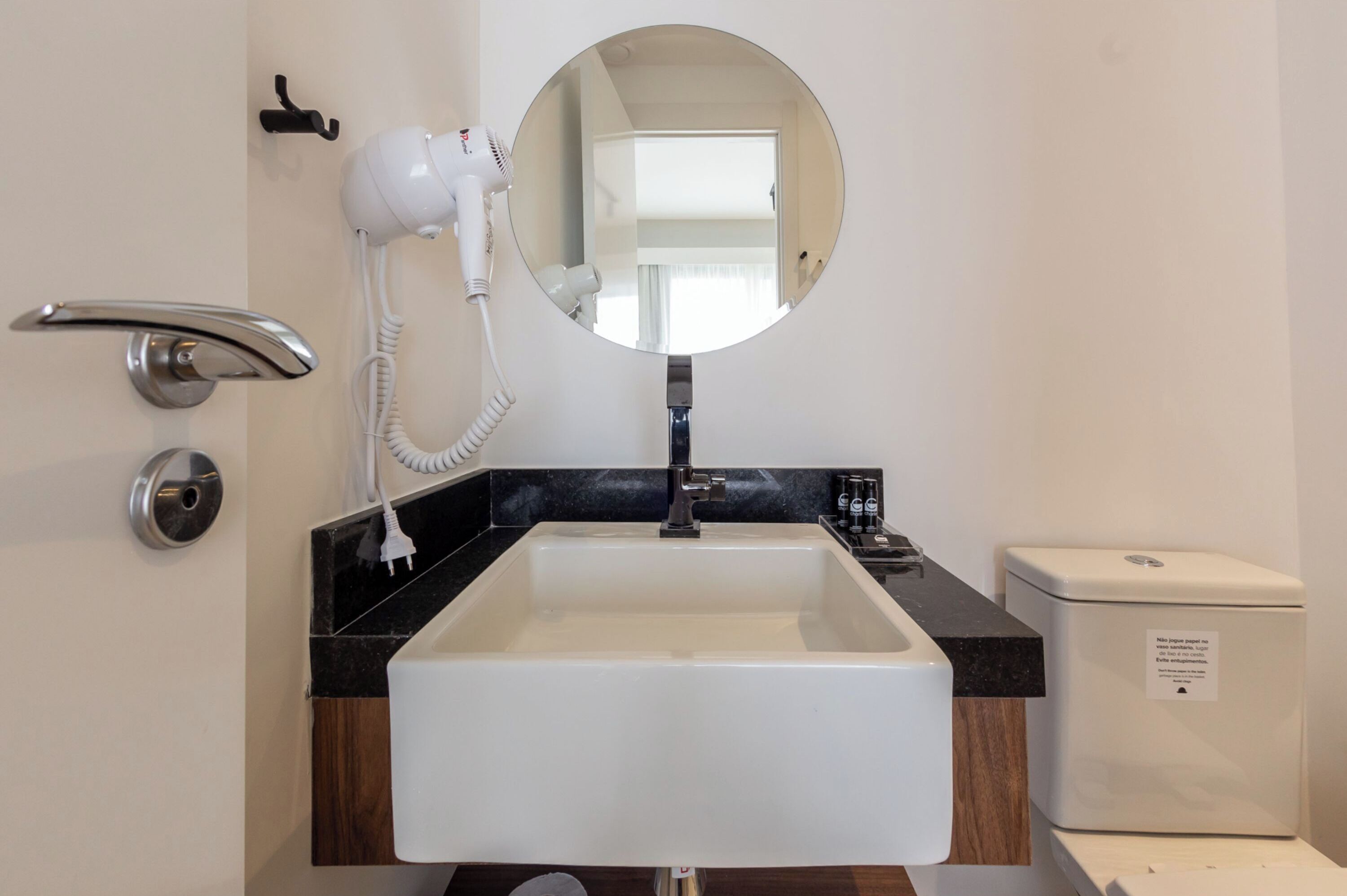 Casa de banho com lavatório branco, espelho redondo, secador de cabelo, sanita e bancada preta.
