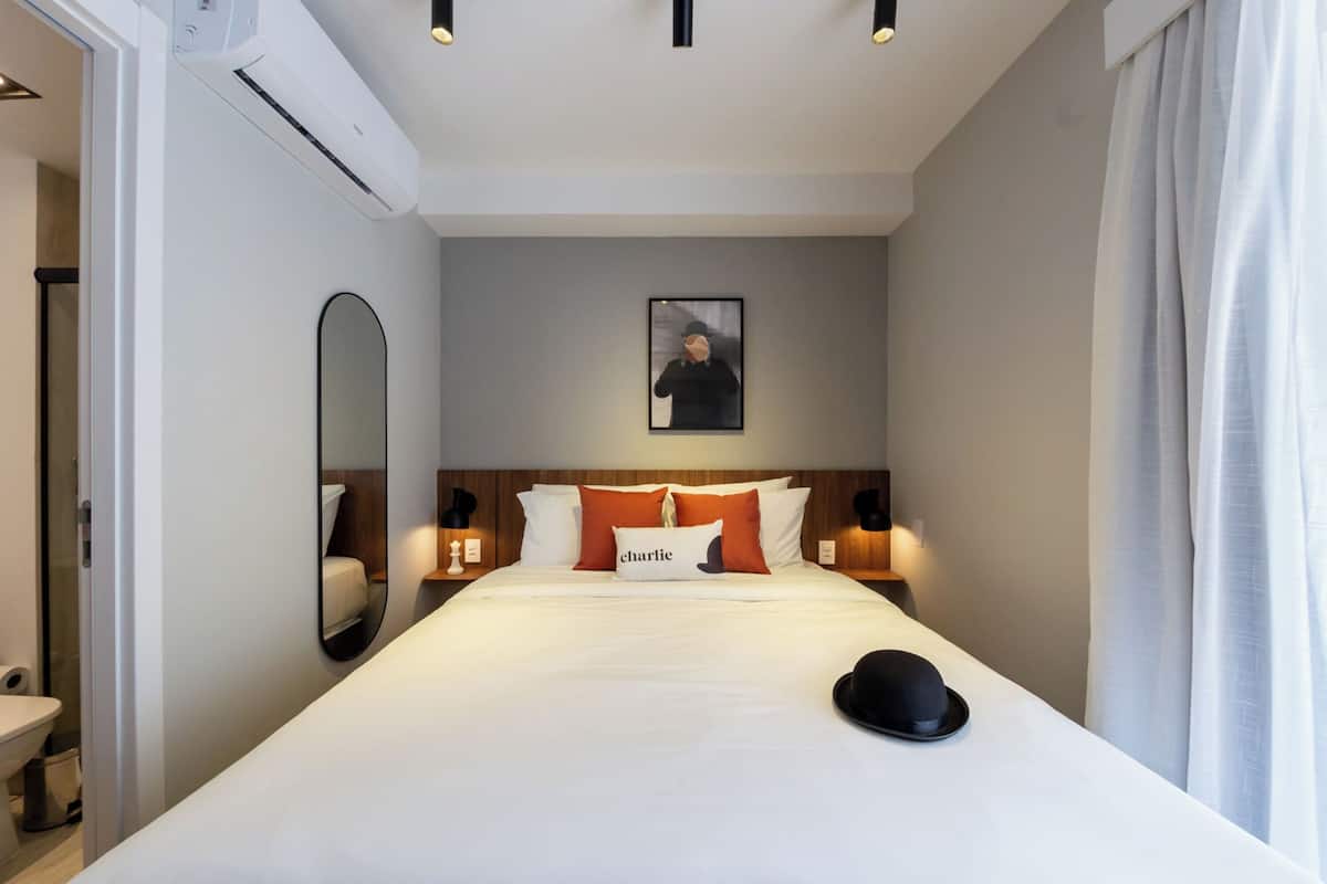 Ein Hotelzimmer mit einem großen Bett, einem Nachttisch, einem Spiegel, einem gerahmten Bild an der Wand und einem Fenster mit Vorhängen.