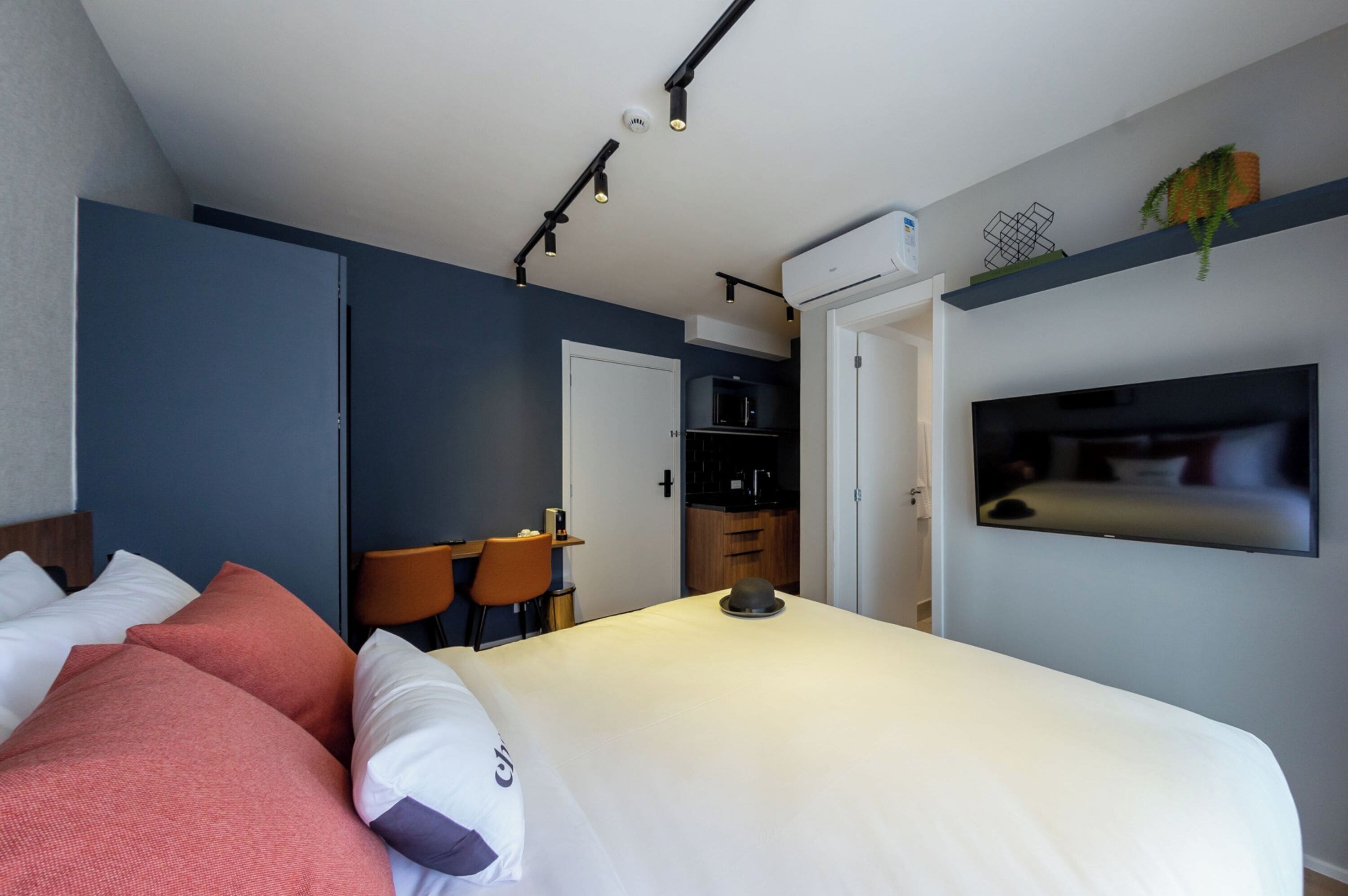 A modern hotel room with a bed, a desk, a chair, a TV, and a kitchenette.