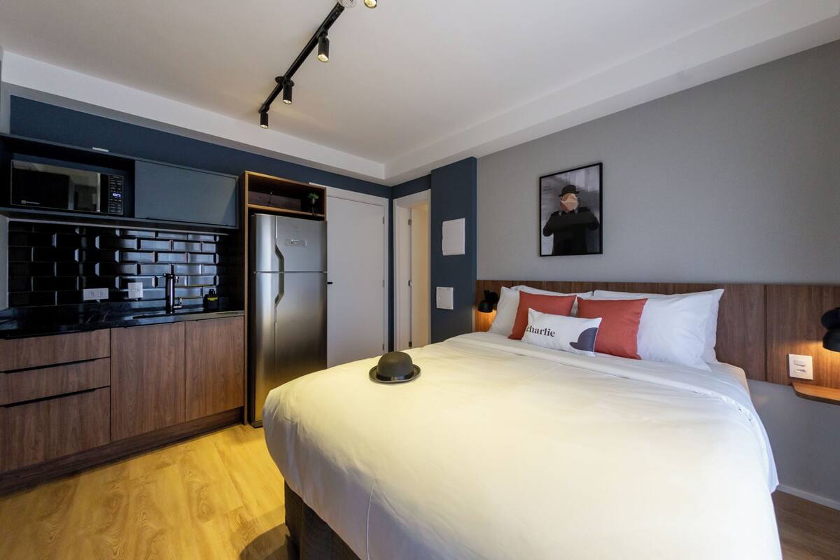 Ein kleines Hotelzimmer mit Küchenzeile, einem Bett mit weißer Bettwäsche und einem Hut, sowie einem gerahmten Bild an der Wand.