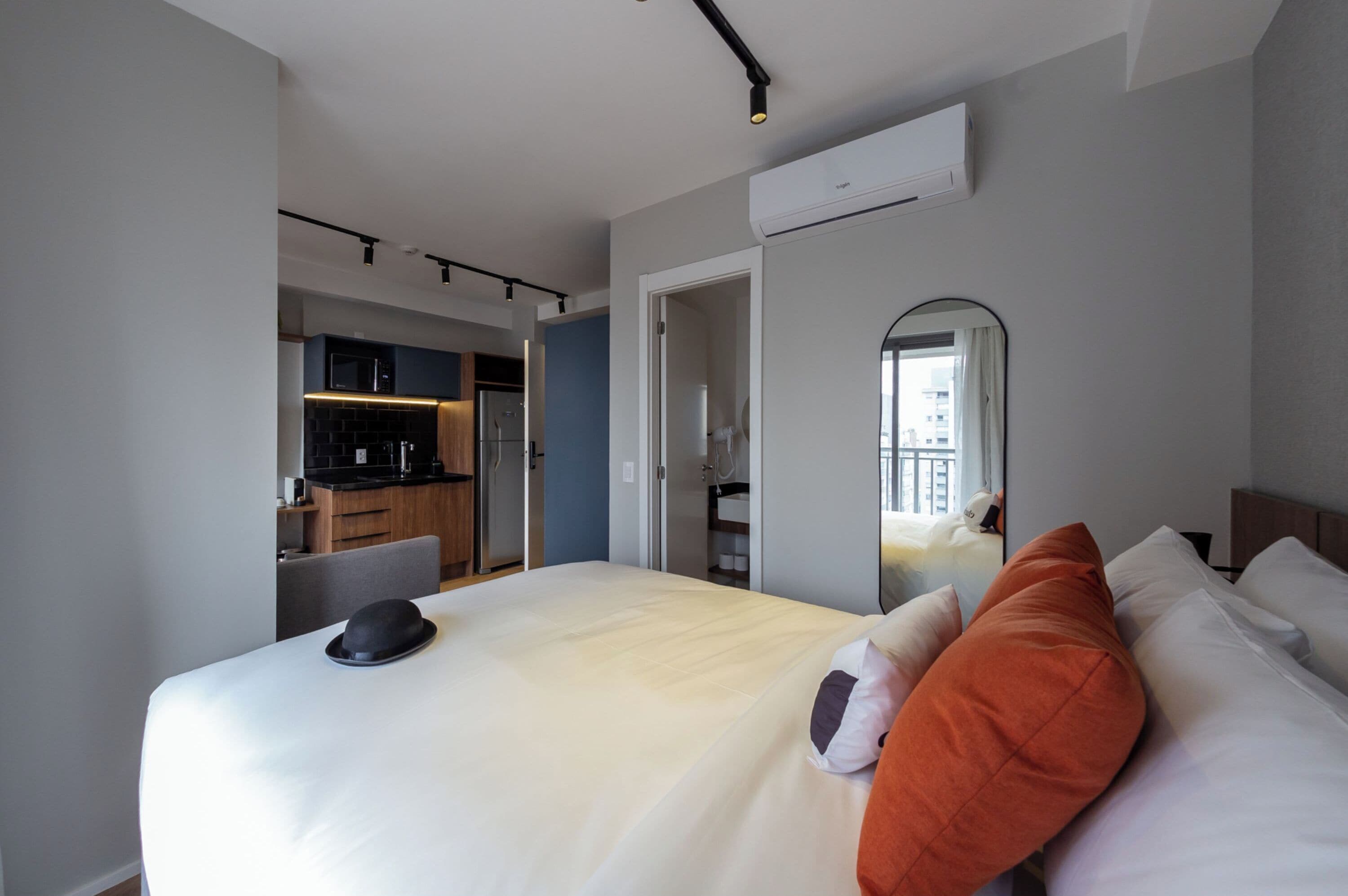 A modern hotel room with a large bed, a small desk, and a kitchenette in the background.