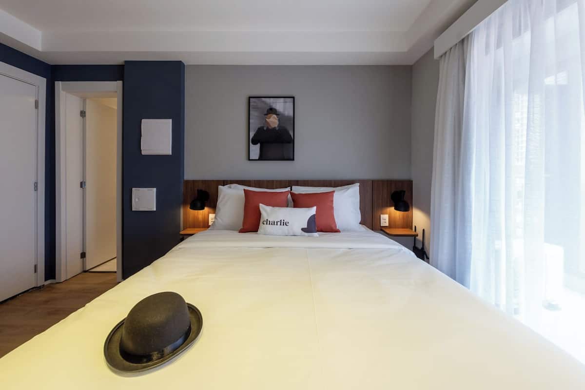 Ein Hotelzimmer mit einem großen Bett, einem Hut auf dem Bett, einem gerahmten Bild an der Wand und einem Fenster mit Vorhängen.