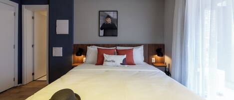 Una habitación de hotel con una cama grande, un sombrero sobre la cama, un cuadro en la pared y una ventana con cortinas.