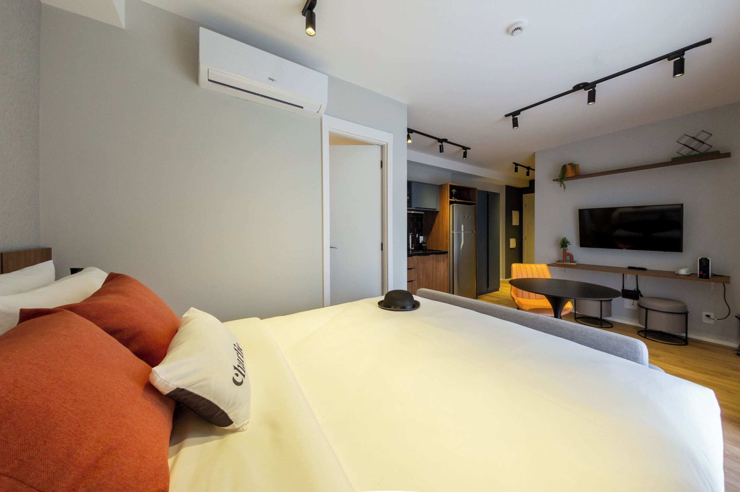 A modern hotel room with a large bed, a dining area with a table and chairs, a TV mounted on the wall, and an air conditioning unit.