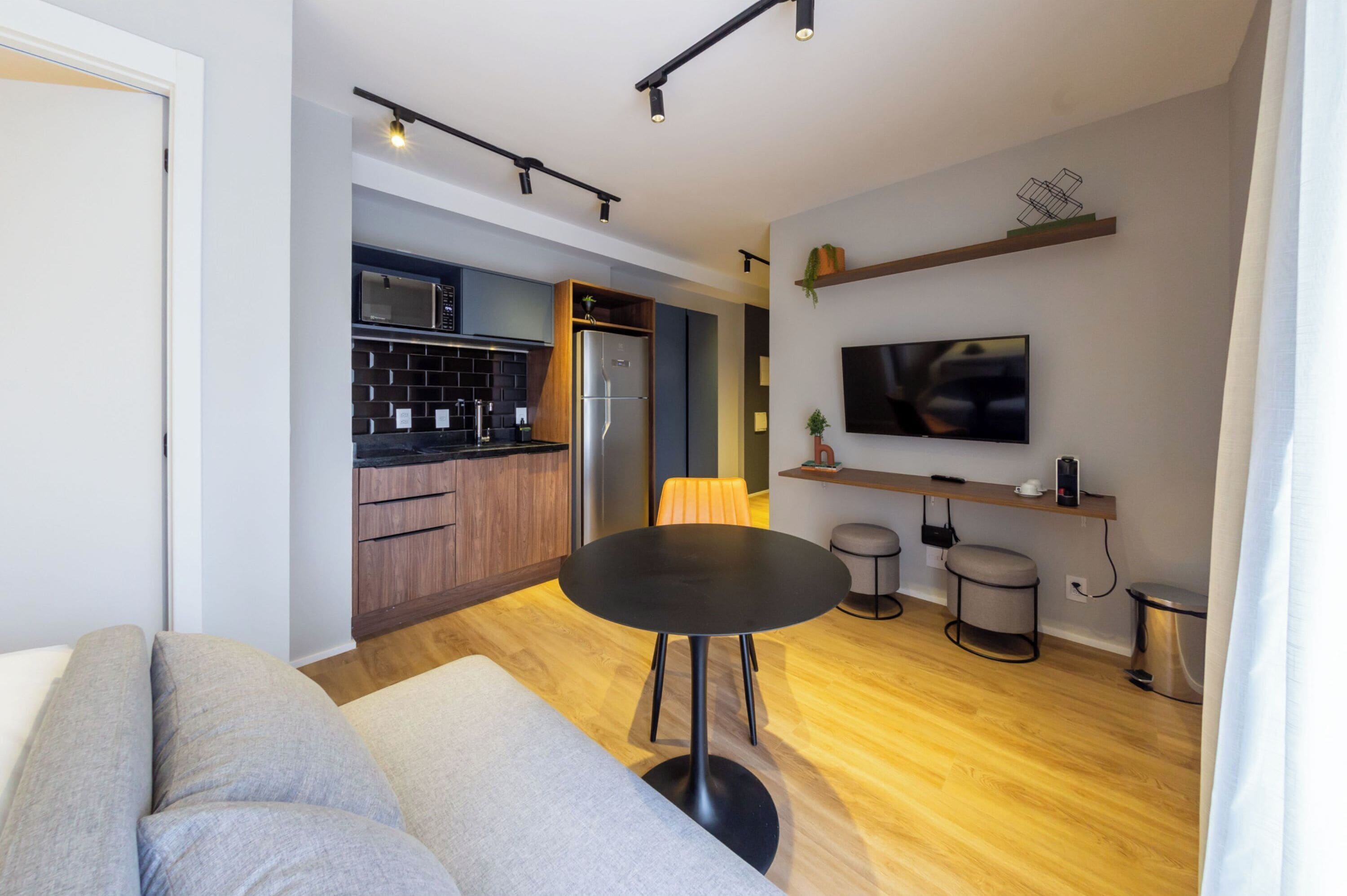 A modern living room with a sofa, a round table, and a kitchen area with a microwave, refrigerator, and wooden cabinets.
