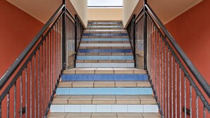 Eine Treppe mit abwechselnd blauen und beigen Stufen, schwarzen Geländern und einem Blick auf den Himmel durch ein Rundbogenfenster oben.