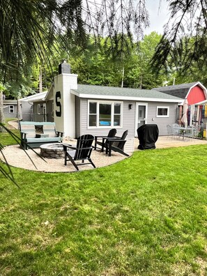 A small house with a covered patio, a grill, and a red barn in the background. - W Lake Mitchell. Priv shared frontage. Snow trail access.Caberfae Peaks,15 miles (Cadillac)