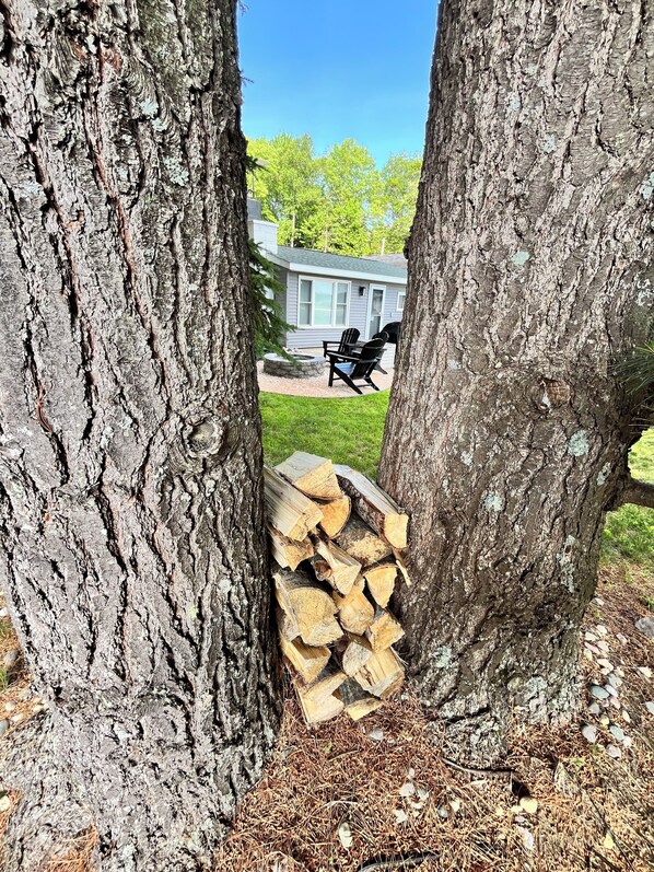 A view through trees to a house with a patio, outdoor seating, and a stack of firewood. - W Lake Mitchell. Priv shared frontage. Snow trail access.Caberfae Peaks,15 miles (Cadillac)