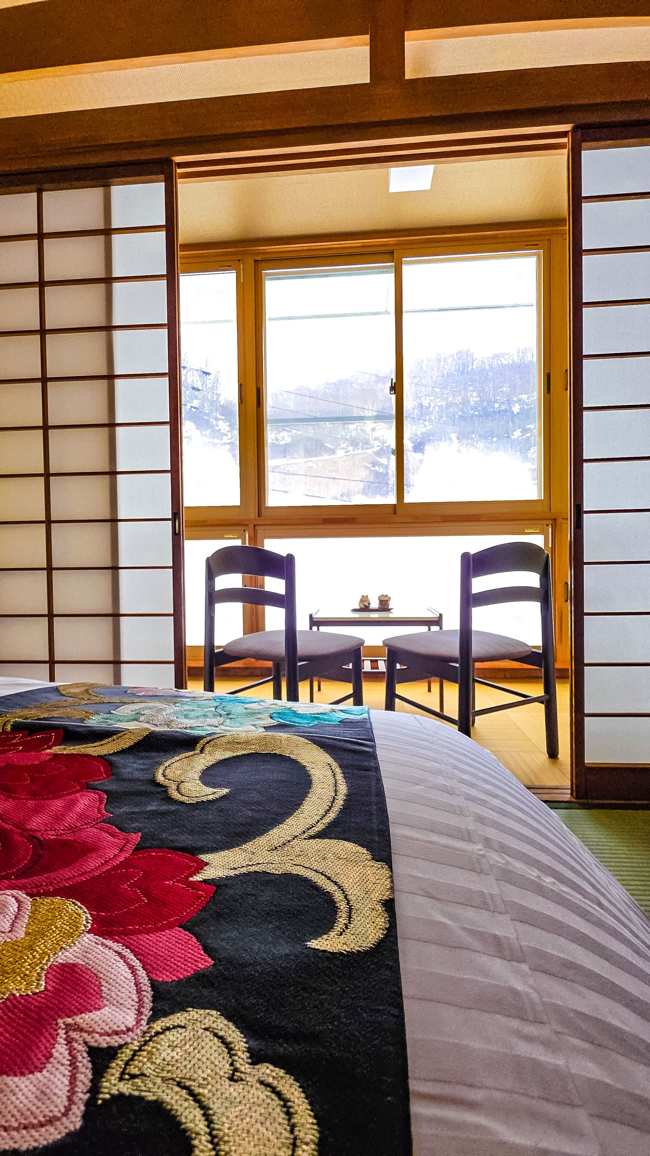 A traditional Japanese room with tatami flooring, a low table with chairs, and a view of a snowy landscape through a large window.