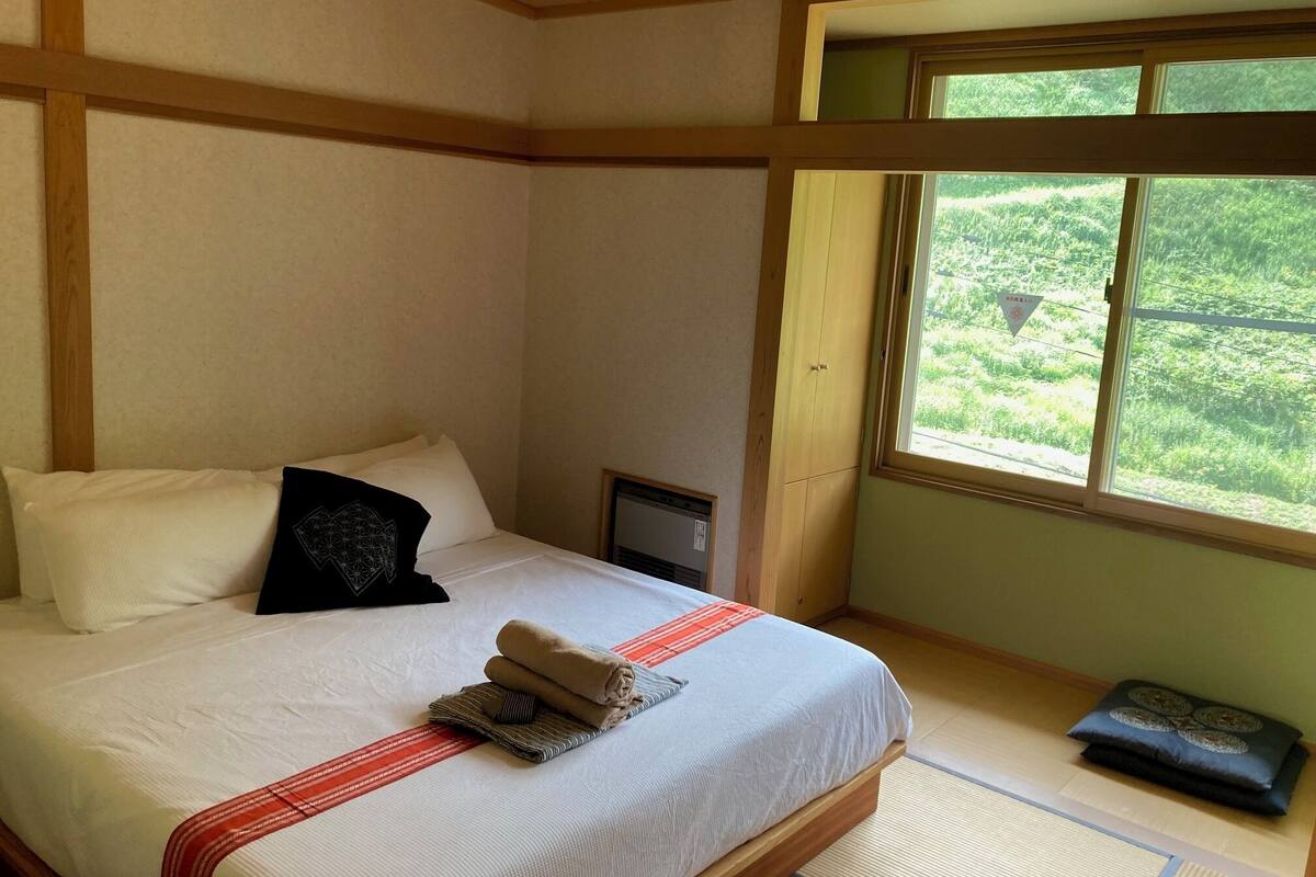 A neatly made bed with pillows, a small heater, and a window with a view of greenery.