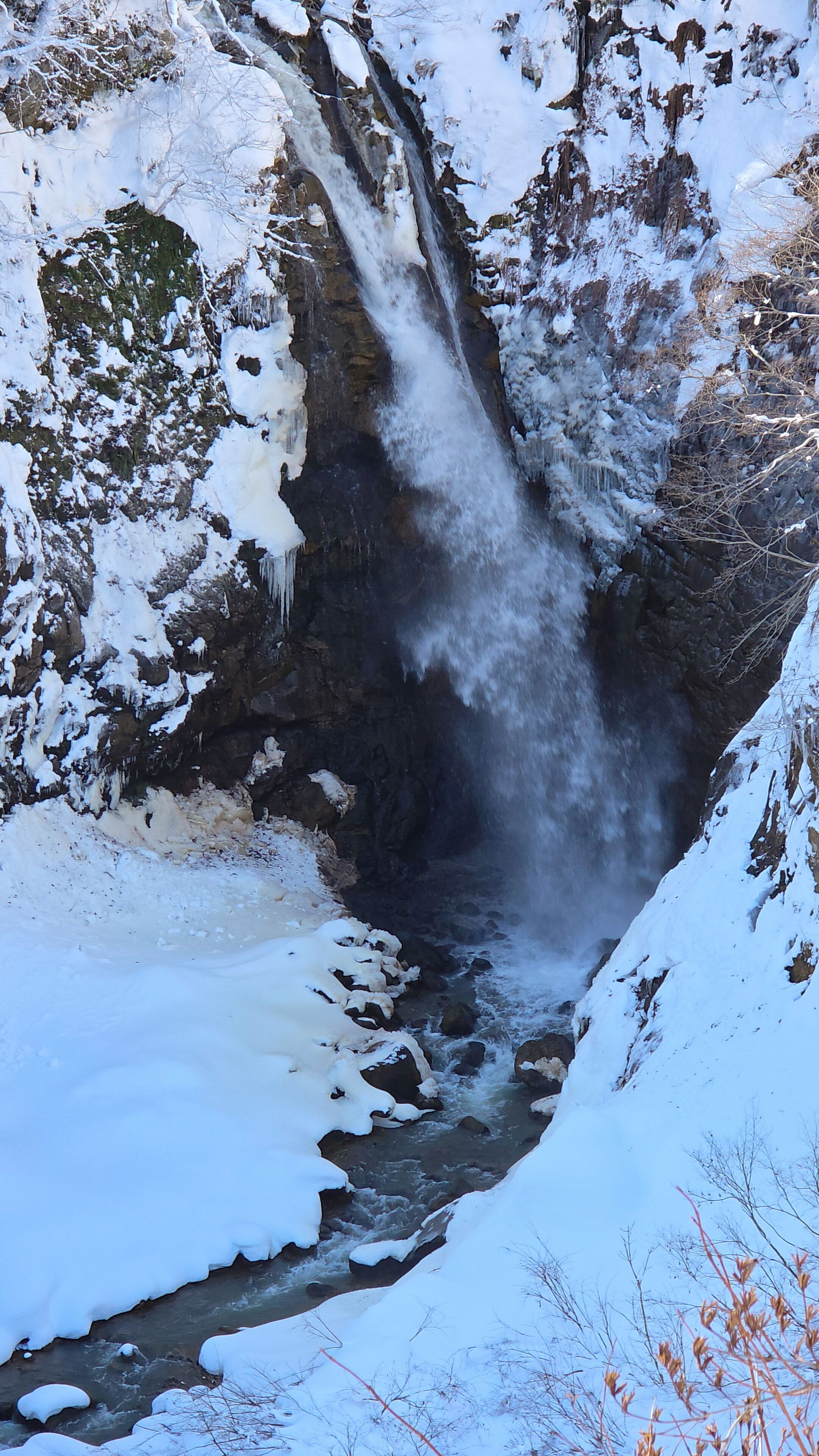 A frozen waterfall surrounded by snow-covered rocks and sparse vegetation.
