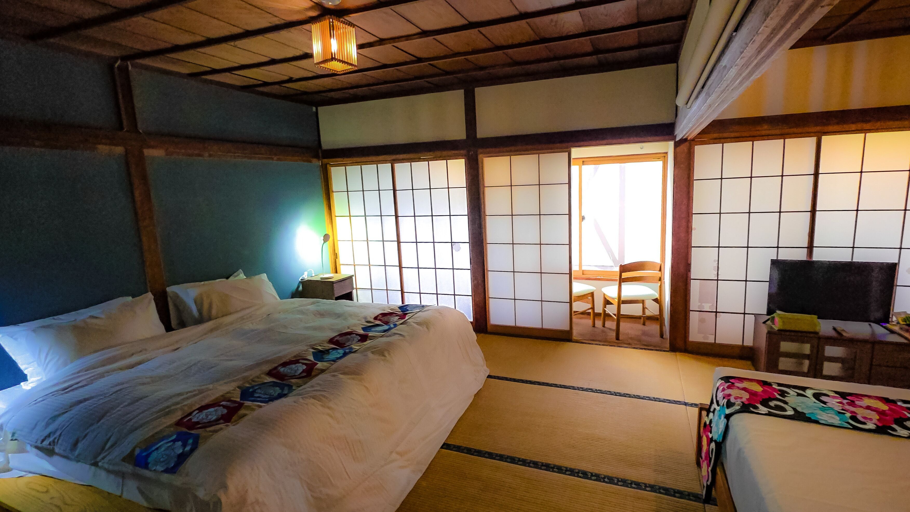 A traditional Japanese-style room with tatami flooring, a bed with a patterned bedspread, a desk with a chair, and sliding doors leading to a balcony.