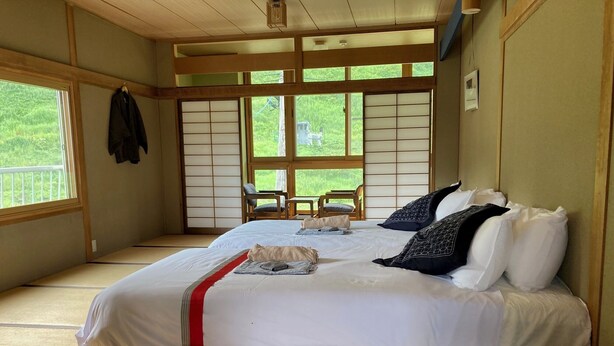 Et traditionelt japansk rum med tatamimåtter, en stor seng med hvide sengetæpper og mønstrede puder, en skydedør af shoji-papir og udsigt til grønne omgivelser.