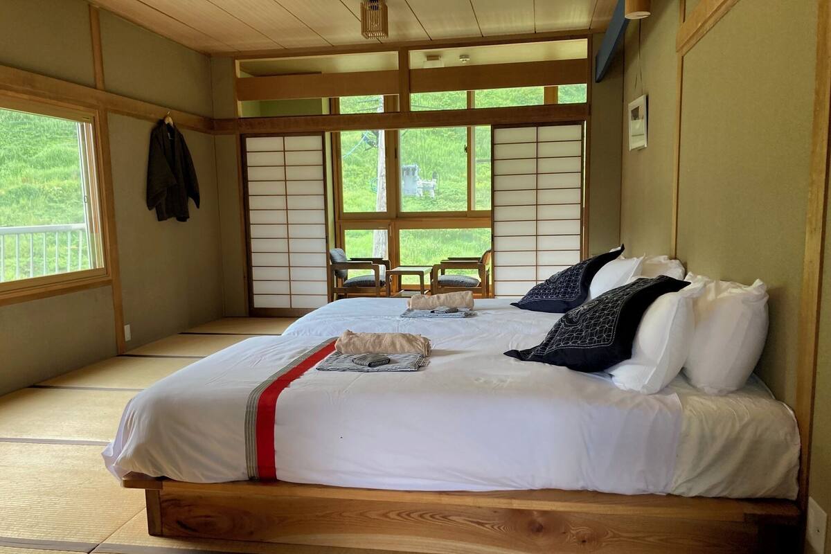A traditional Japanese-style room with tatami flooring, a large bed with white bedding and patterned pillows, a sliding shoji screen door, and a view of greenery outside.