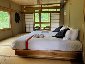 Una habitación tradicional japonesa con suelo de tatami, una cama grande con ropa de cama blanca y almohadas estampadas, una puerta corrediza de shoji y vistas a la vegetación exterior.