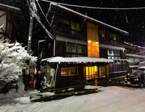 A building with a snow-covered roof and a lit window, surrounded by snow. - Komatsuya Seki Onsen (Myoko)