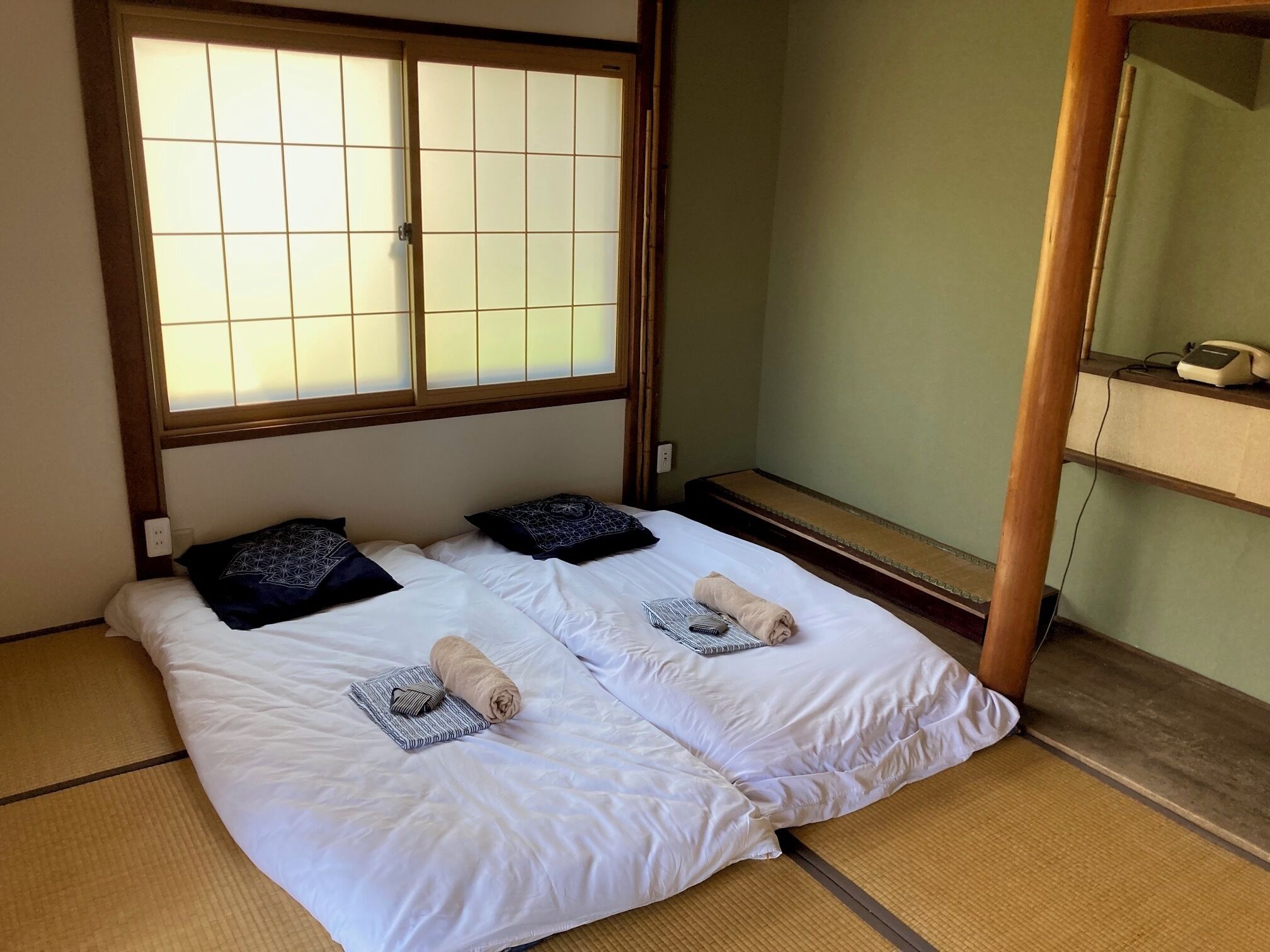 A traditional Japanese-style room with tatami flooring, a shoji screen window, and a bed with two pillows and a rolled towel.