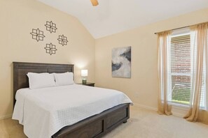 A bedroom with a large bed, a nightstand with a lamp, a wall art piece, and a window with curtains. - Spacious Home. Pool. Hot Tub. Grill. Pool Table. 4 Bed. 2 Bath. (Watauga)