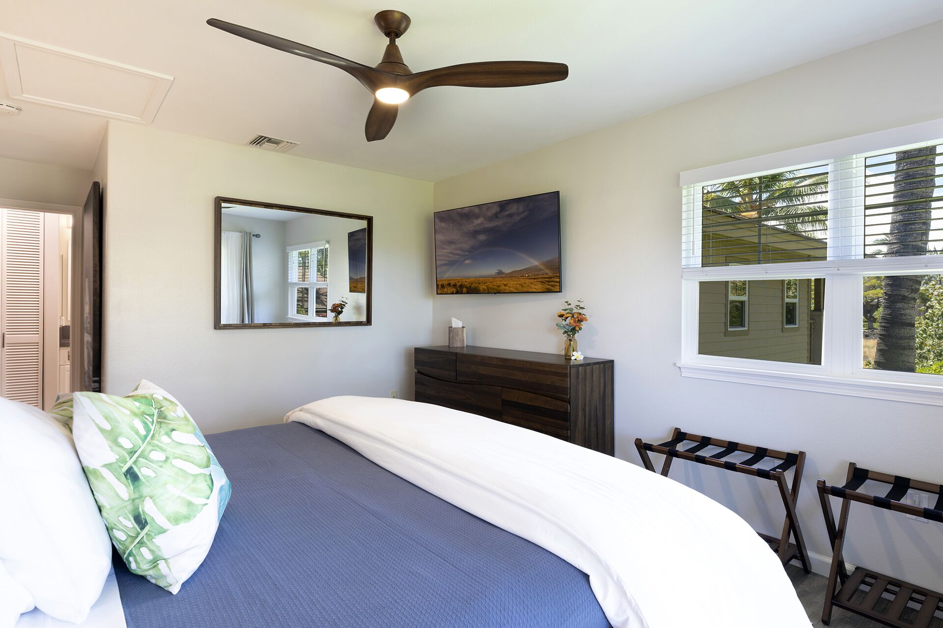 Une chambre à coucher comprenant un lit, une commode, un miroir, un ventilateur de plafond, une fenêtre et une vue sur les arbres.