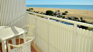 Un balcón con vista a una playa, mobiliario blanco y una valla blanca.