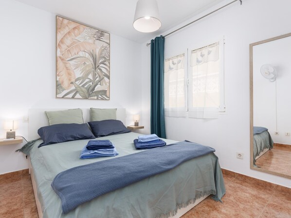 A neatly made bed with a blue and green bedspread, two pillows, and folded towels. There is a large mirror on the wall, a ceiling light, and a window with curtains. - Casa Marcelo, charming and renovated house (Antigua)