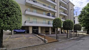 Un immeuble résidentiel à plusieurs étages, doté d’un espace de stationnement et d’arbres soigneusement taillés. Une voiture bleue est garée devant.