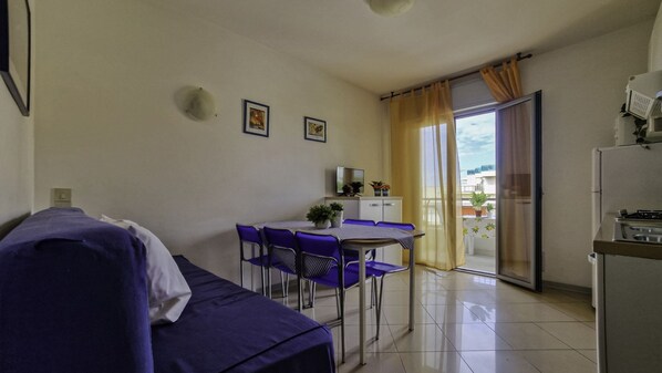 Une pièce compacte comprenant un lit, une petite table à manger avec des chaises, un coin cuisine et une porte-fenêtre coulissante donnant sur un balcon.