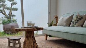Ein modernes Wohnzimmer mit einem hellgrünen Sofa, einem Holztisch und einem kleinen Holzschemel. Eine große Topfpflanze und Blick auf Bäume draußen.
