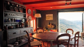 A dining area with a round table and chairs, a wooden cabinet with dishes, and a view of mountains through a large window. - Chalet mountain and dog friendly, hot tub (Bourg-Saint-Maurice)