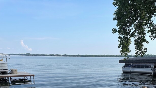 Dos sillones Adirondack de madera mirando a un lago tranquilo con un muelle y un bote cubierto al fondo.