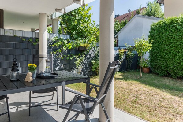 An outdoor patio with a table and chairs, surrounded by greenery and a house in the background.