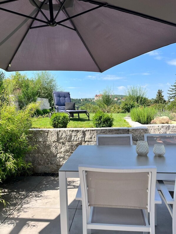 An outdoor patio with a table and chairs, a large umbrella, and a view of a garden and distant buildings. - Holiday Apartment 'Burgblick' with Private Terrace, Garden & Wi-Fi (Harburg)