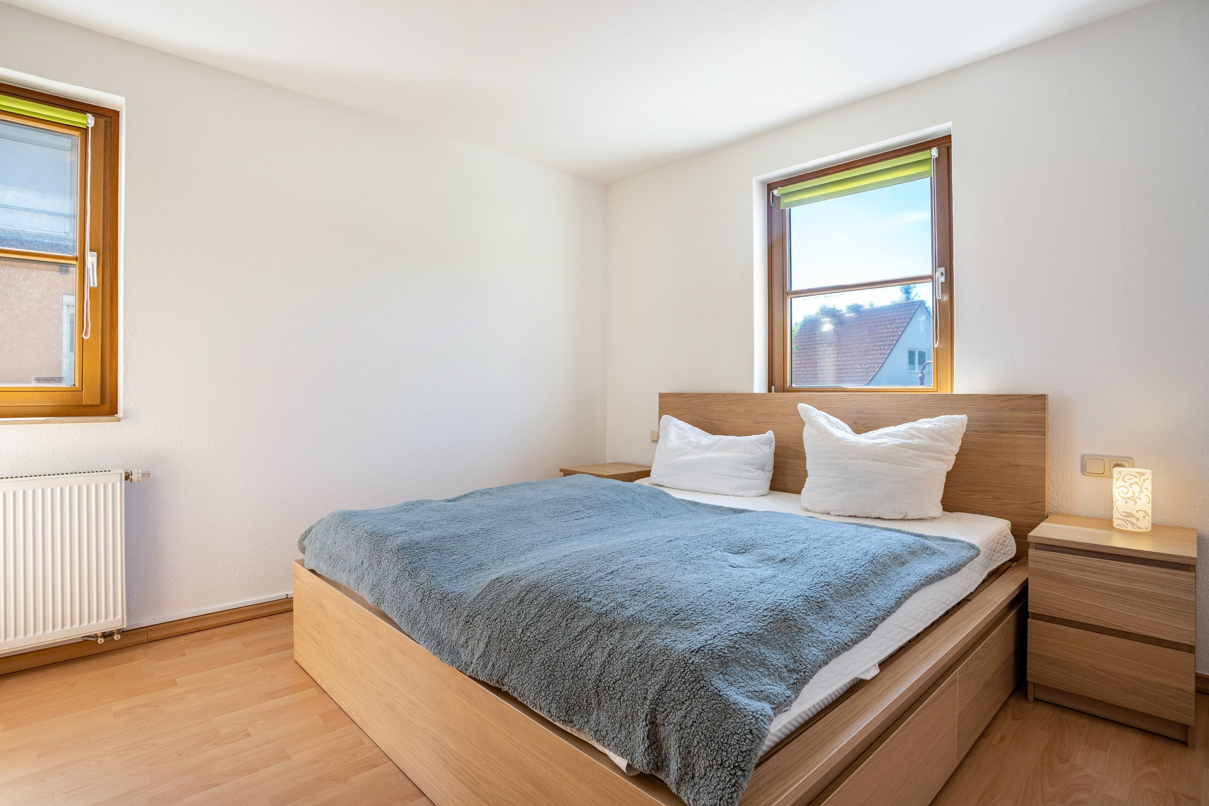 Une chambre avec un lit en bois, des oreillers blancs, une couverture bleue, une table de chevet avec une lampe et une fenêtre donnant sur un beau paysage.
