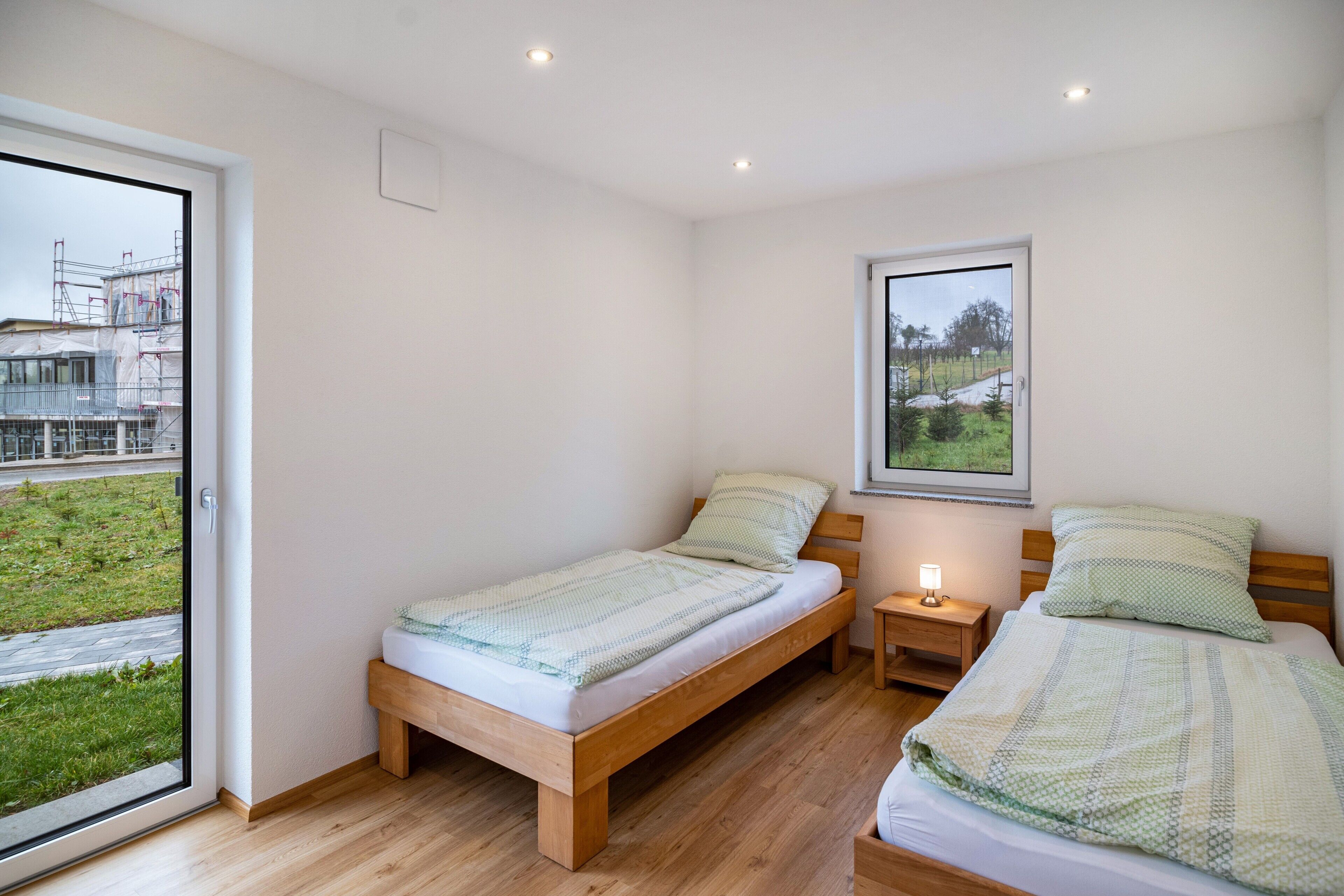 A bedroom with two beds, a window with a view of a building under construction, and a nightstand with a lamp.
