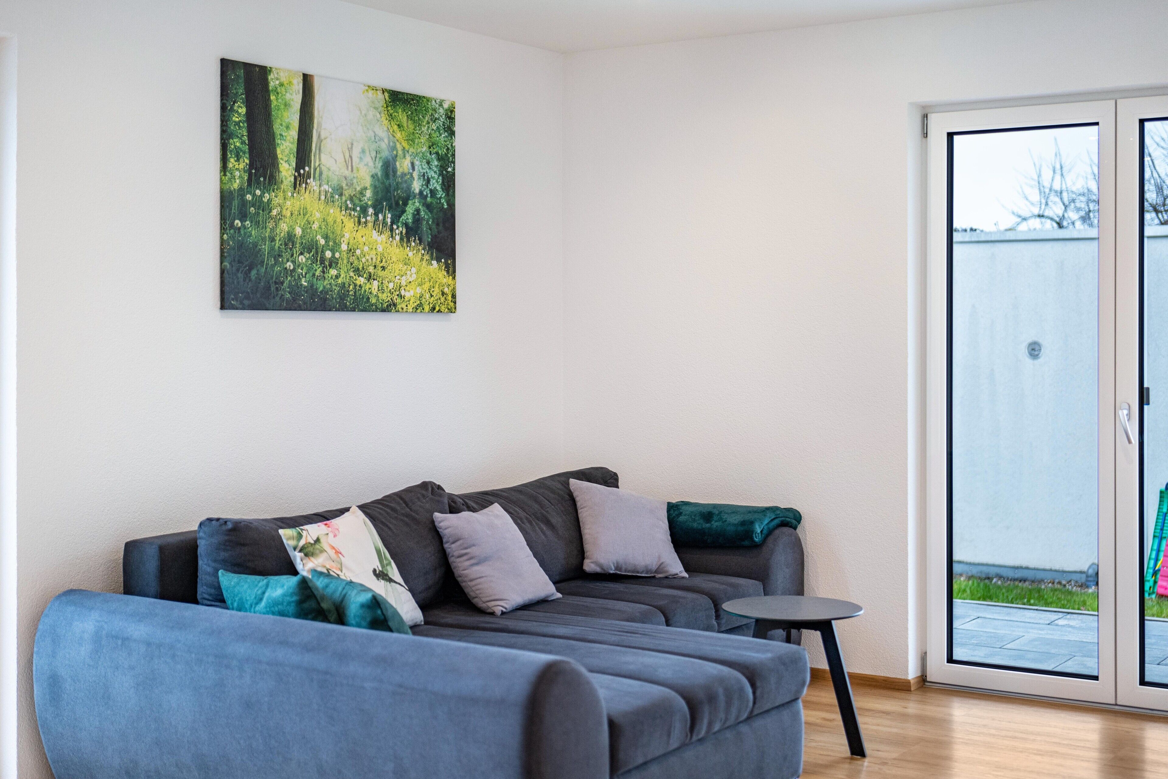 Ein modernes Wohnzimmer mit einem grauen Sofa, einem kleinen runden Tisch und einer Schiebetür zu einem Außenbereich.